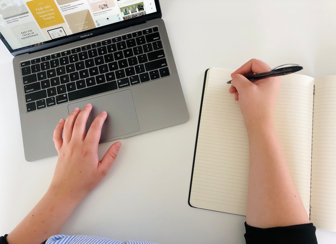 Hand typing on a laptop keyboard while writing notes in a notebook, planning LinkedIn content