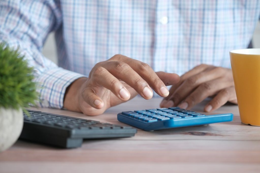 Hand using a calculator next to a coffee mug while working out cost-per-meeting outreach numbers at a desk